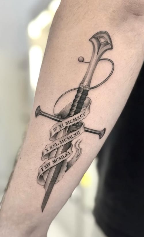 A black and grey tattoo of a realistic sword with Roman numerals on the forearm, featuring detailed shading and script elements — by otaybeatdown, los angeles