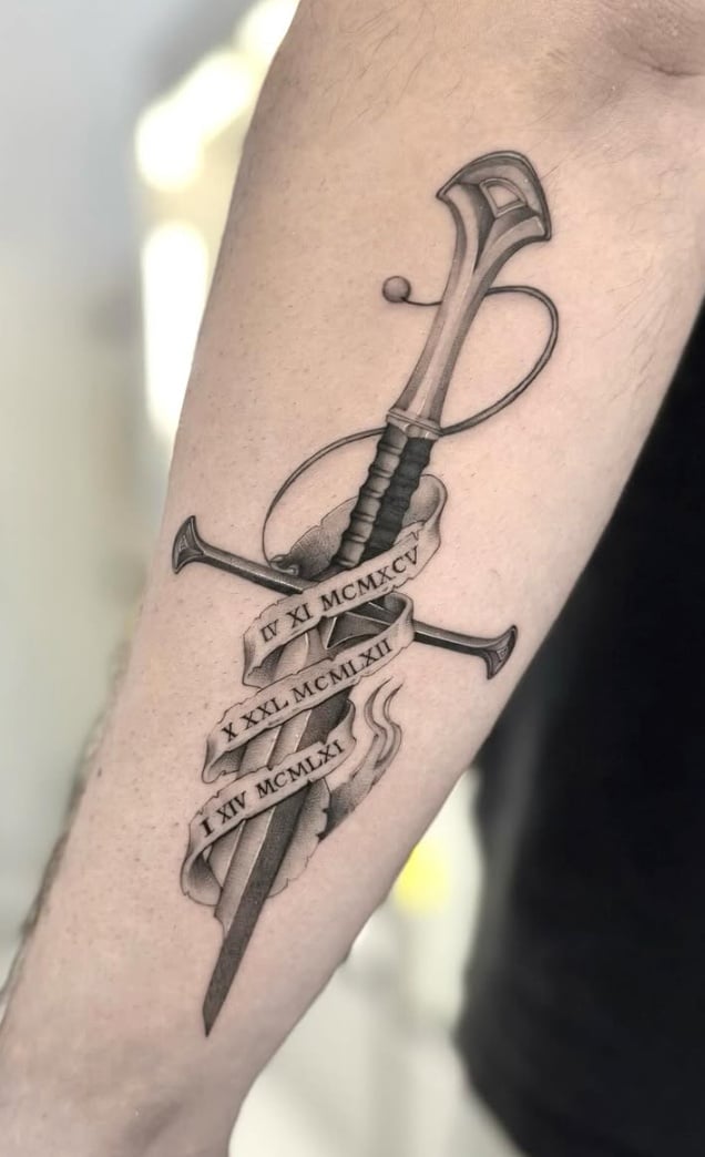 A black and grey tattoo of a realistic sword with Roman numerals on the forearm, featuring detailed shading and script elements — by otaybeatdown, los angeles