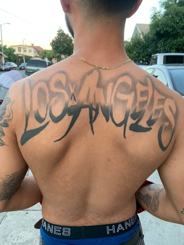 A bold black and grey lettering tattoo of 'Los Angeles' across the upper back with stylized script and sharp edges — by Humilde tattoos, los angeles