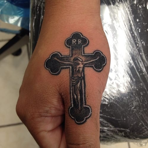A bold black and grey crucifix tattoo on the hand featuring Jesus Christ, with a strong religious theme — by Johnny Ayala, los angeles