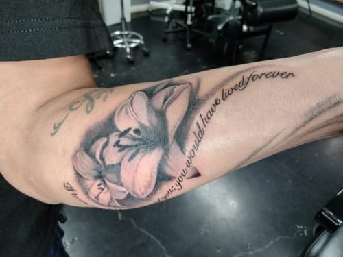 A black and grey tattoo on the forearm featuring a realistic flower with accompanying script text — by anoetic.designs.ultd, los angeles