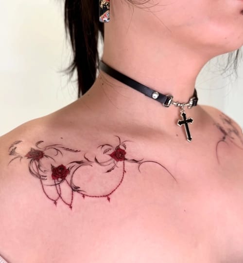 A delicate fine line tattoo on the collarbone featuring ornamental swirls and red roses, combining black and red ink for a striking effect — by zitronxus, los angeles