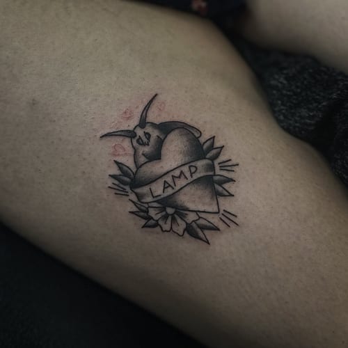 A black and grey tattoo of a moth with a heart and banner on the thigh — by Maddie Ramirez, los angeles