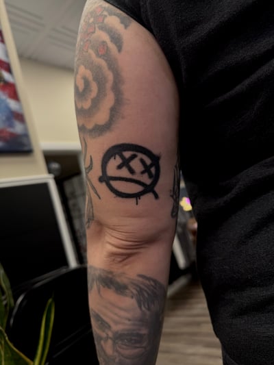 A minimalist blackwork tattoo of a face with crossed-out eyes and a dripping mouth on the upper arm — by emily, los angeles