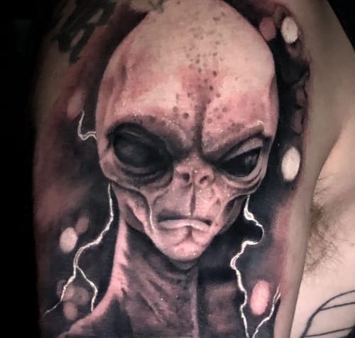 A black and grey realism tattoo of an alien head on the upper arm, featuring detailed shading and a dark, mysterious atmosphere — by Travis Ross, los angeles