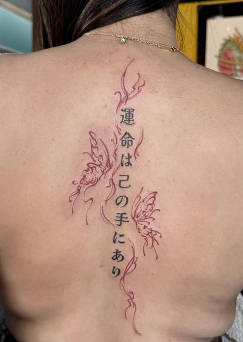 A fine line tattoo on the back featuring delicate butterflies and Japanese script in red and black ink — by Shelby Bianco, los angeles