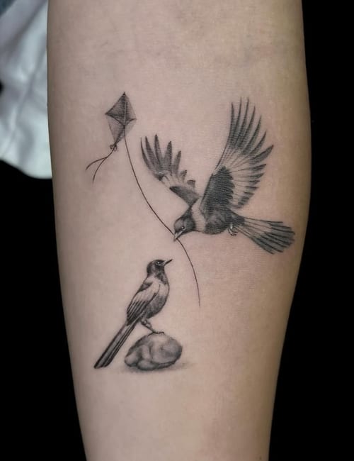 A black and grey fine line tattoo of two birds, one flying with a kite string and the other perched on a rock, located on the forearm — by Jian Liang, los angeles