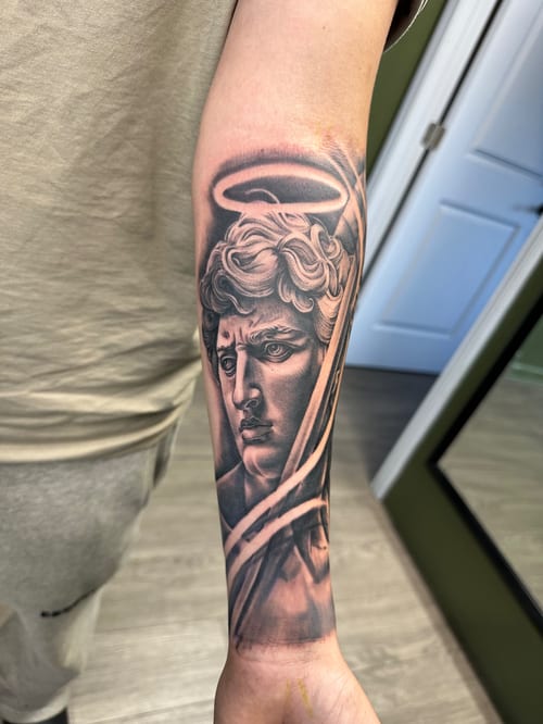 A black and grey realism tattoo of Michelangelo's David on the forearm, featuring detailed shading and a halo above the head — by Humilde tattoos, los angeles