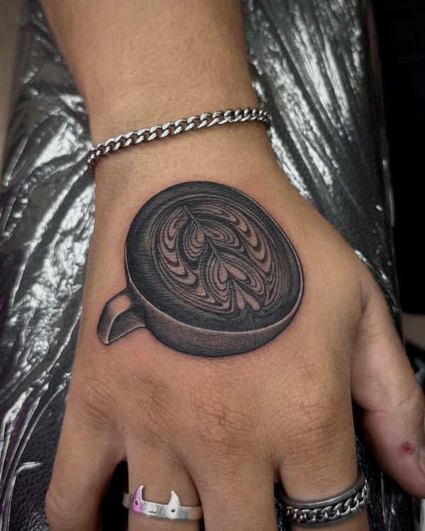 A realistic black and grey tattoo of a coffee cup with intricate latte art on the hand — by emily, rahway