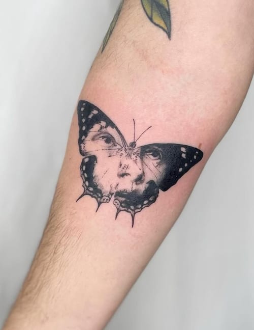 A surreal black and grey tattoo of a butterfly with a human face on the forearm, combining intricate details and bold outlines — by Level tattoo, los angeles