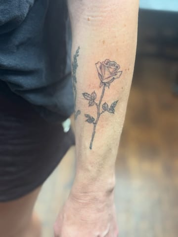 A delicate fine line rose tattoo on the forearm, featuring minimalistic black and grey shading — by Sandra Price, los angeles