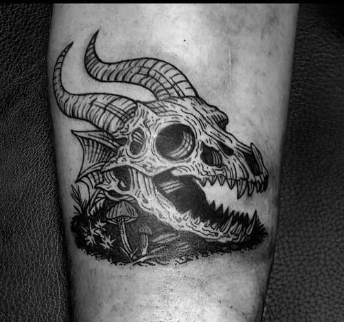 A black and grey tattoo of a horned skull with mushrooms growing around it, located on the forearm. The design features intricate linework and shading, giving it a dark and mysterious appearance — …
