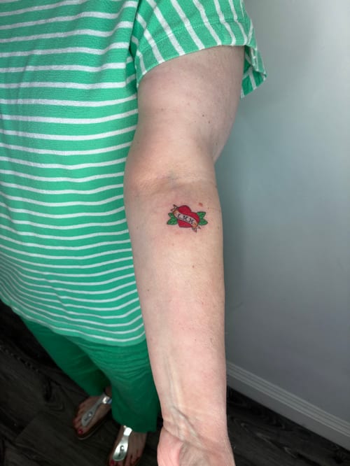 A small traditional style tattoo of a red heart with a banner on the forearm, featuring bold colors and simple lines — by Edgar Armas Rodriguez, los angeles