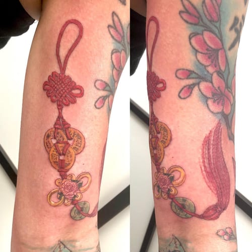 A colorful traditional style tattoo of a Chinese knot with coins and flowers on the forearm — by Sandra Price, los angeles
