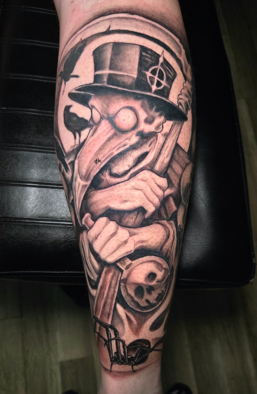 A black and grey Neo-Traditional tattoo on the calf depicting a plague doctor with a top hat, surrounded by crows and skulls, featuring bold lines and dark shading — by Dretty, los angeles