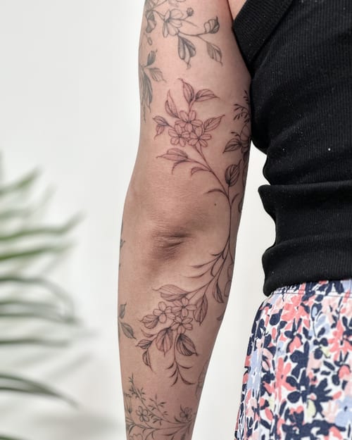 A delicate fine line floral tattoo on the upper arm, featuring intricate black and grey flowers and leaves — by Melanie Benoit, los angeles