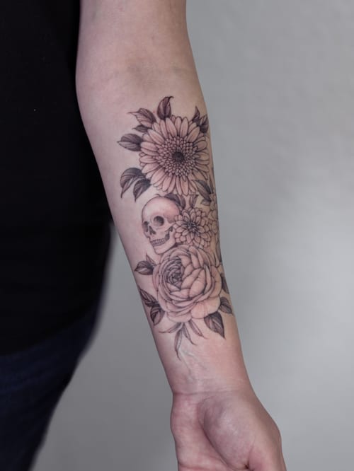 A delicate black and grey tattoo of a skull surrounded by flowers on the forearm — by Melanie Benoit, los angeles