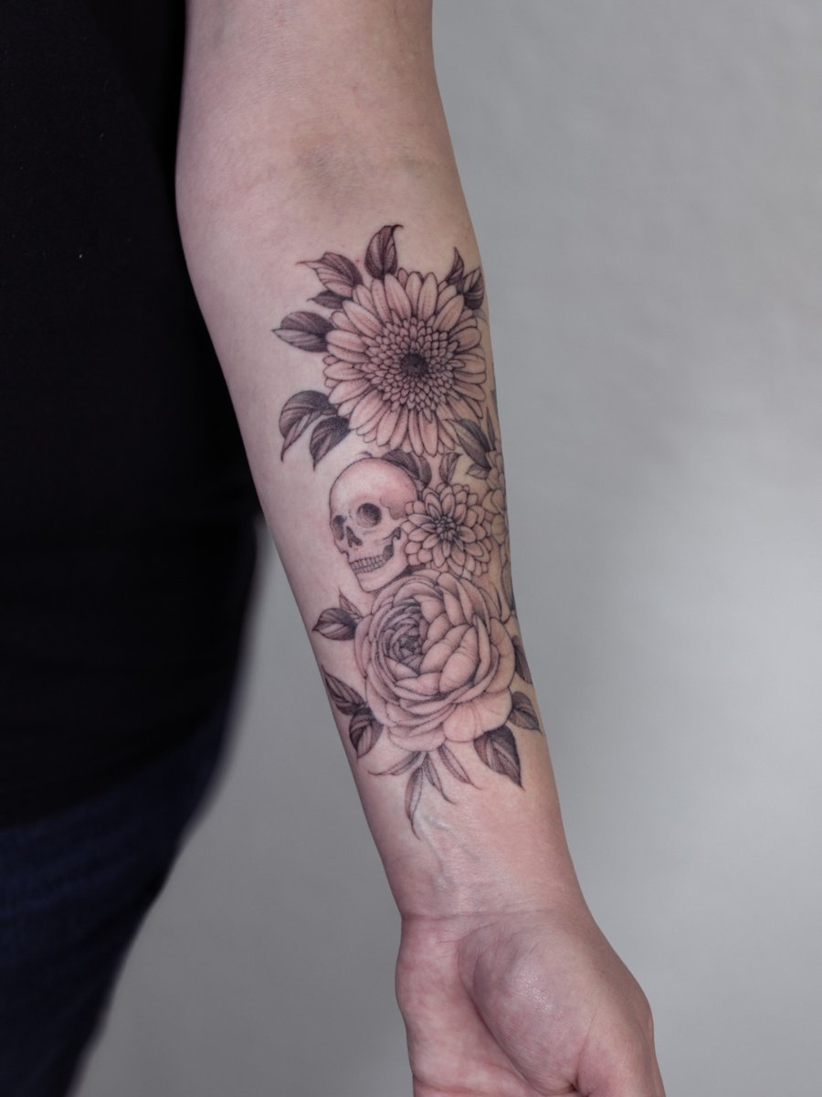 A delicate black and grey tattoo of a skull surrounded by flowers on the forearm — by Melanie Benoit, los angeles