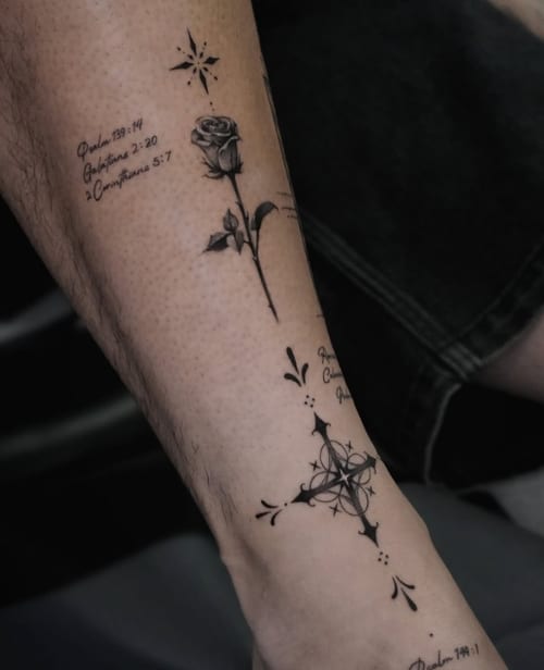 A fine line black and grey tattoo on the forearm featuring a rose, script, and a decorative cross with delicate linework — by Jian Liang, los angeles