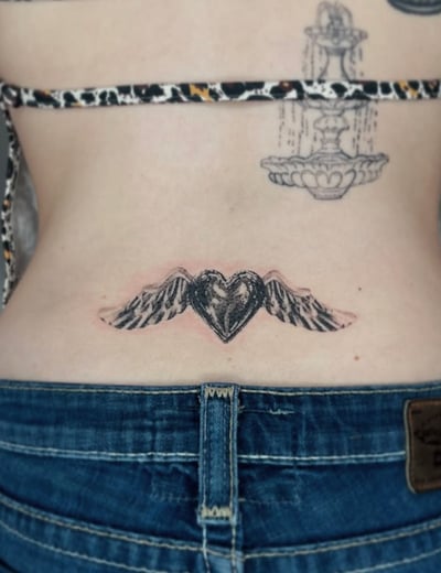 A black and grey tattoo of a heart with wings on the lower back, featuring bold outlines and shading — by Level tattoo, los angeles
