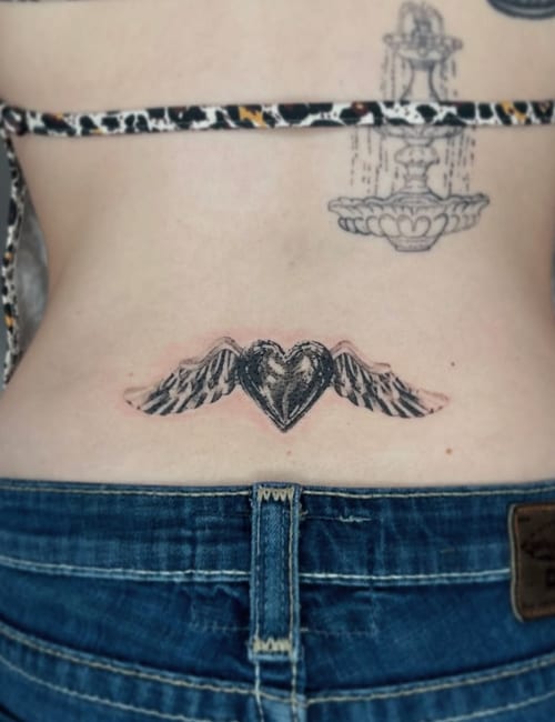 A black and grey tattoo of a heart with wings on the lower back, featuring bold outlines and shading — by Level tattoo, los angeles