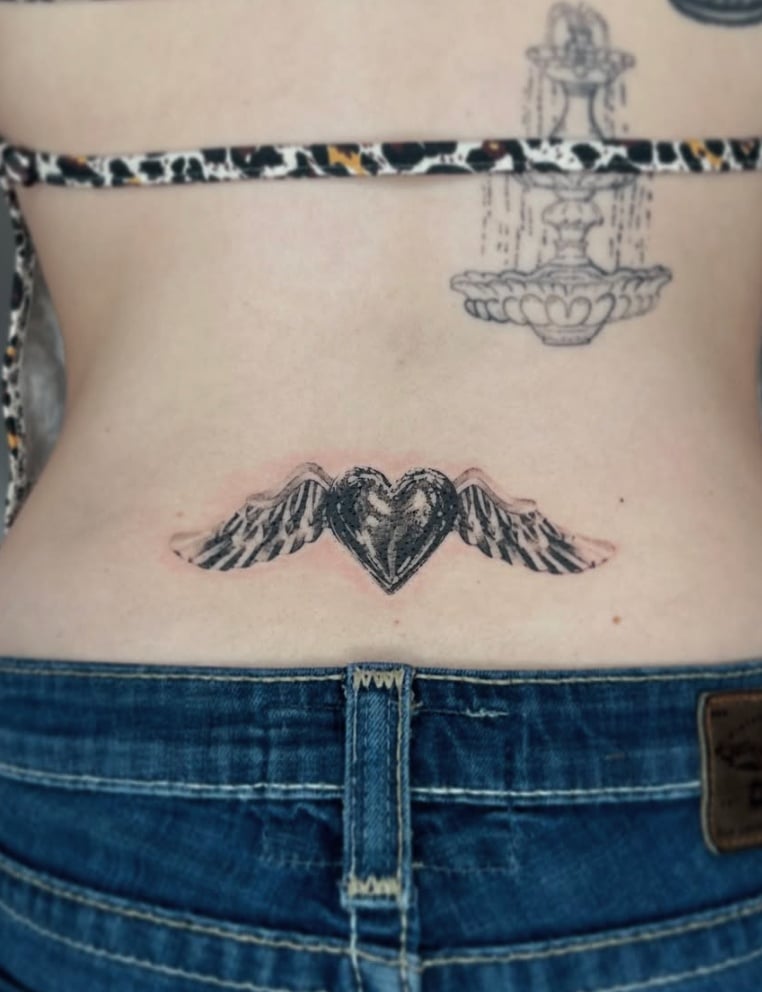A black and grey tattoo of a heart with wings on the lower back, featuring bold outlines and shading — by Level tattoo, los angeles
