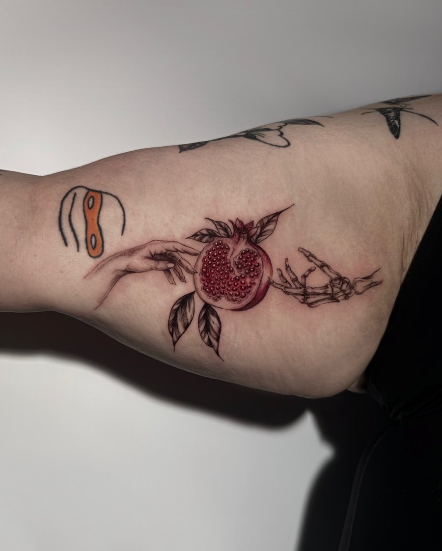 A delicate illustrative tattoo of a pomegranate held by a human and skeleton hand on the arm, featuring fine line work and mixed color — by Shelby Bianco, los angeles