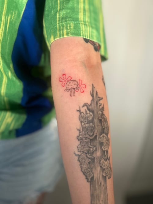 A small, whimsical jellyfish tattoo on the forearm with minimalist linework and red accents — by Brian Kies, los angeles