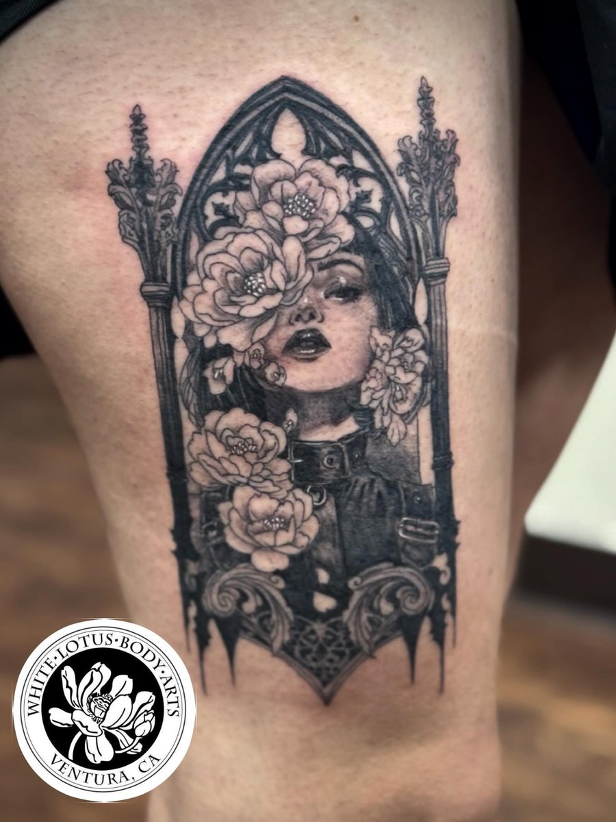 A black and grey tattoo of a woman's portrait surrounded by flowers on the thigh, featuring ornate and delicate details — by Sandra Price, los angeles