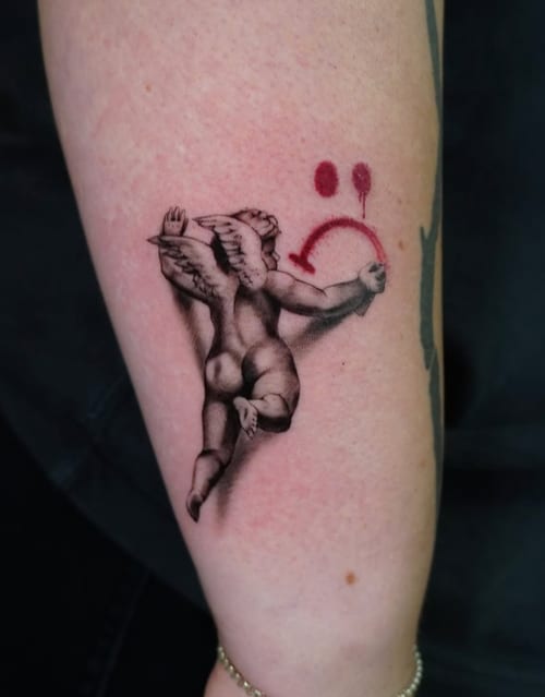 A black and grey illustrative tattoo of an angel on the forearm, with a red smiley face element adding a pop of color — by Jian Liang, los angeles