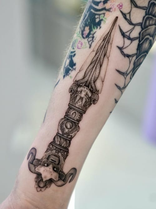 A black and grey realism tattoo of an ornate dagger on the forearm, featuring intricate details and shading — by otaybeatdown, los angeles