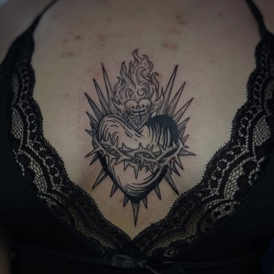 A bold black and grey sacred heart tattoo on the sternum with flames and thorns — by Maddie Ramirez, los angeles