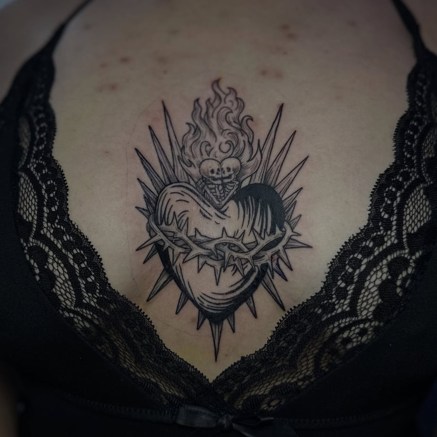 A bold black and grey sacred heart tattoo on the sternum with flames and thorns — by Maddie Ramirez, los angeles