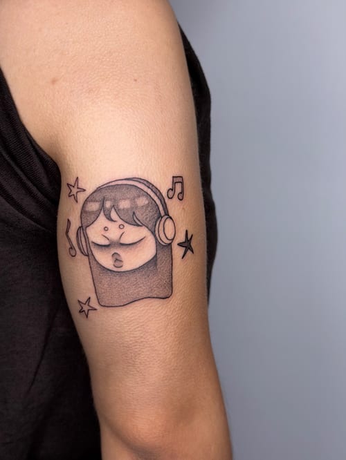A minimalist black and grey tattoo of a cartoon face wearing headphones, surrounded by musical notes and stars, located on the upper arm — by lunayflorestattoo, los angeles