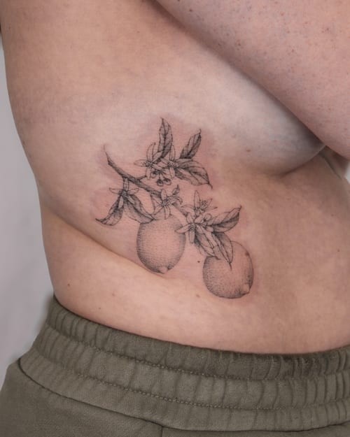 A delicate black and grey fine line tattoo of lemons with leaves on the rib, featuring botanical details — by Melanie Benoit, los angeles