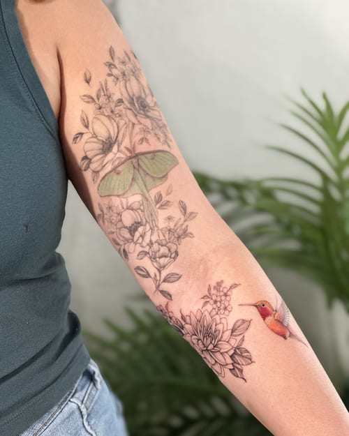A delicate tattoo of a colorful hummingbird with fine line flowers on the forearm, blending realism and fine line styles — by Melanie Benoit, los angeles