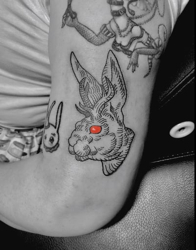A linework tattoo of a jackalope with red eyes on the upper arm, featuring bold black outlines and minimal color accents — by Tag Devilish, los angeles