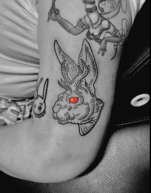 A linework tattoo of a jackalope with red eyes on the upper arm, featuring bold black outlines and minimal color accents — by Tag Devilish, los angeles