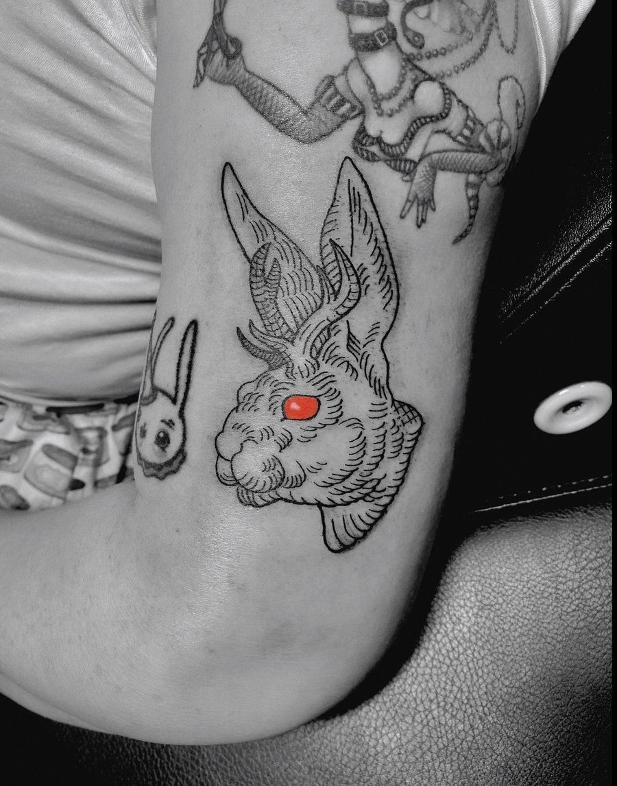 A linework tattoo of a jackalope with red eyes on the upper arm, featuring bold black outlines and minimal color accents — by Tag Devilish, los angeles
