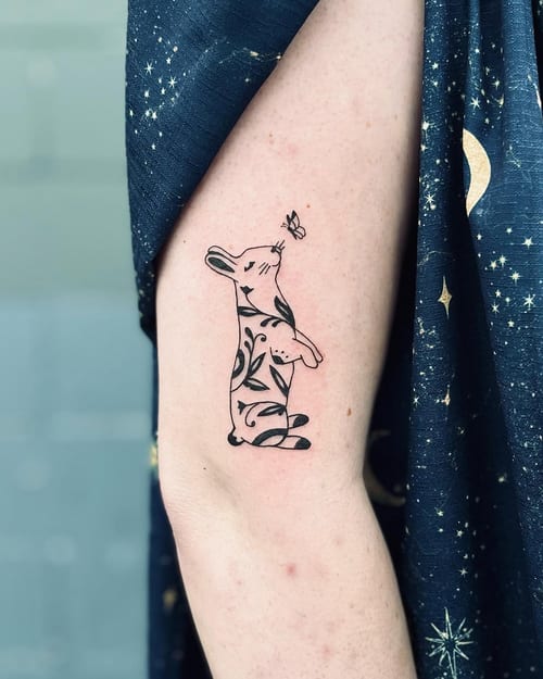 A whimsical minimalist tattoo of a rabbit with a butterfly on the arm, featuring black and grey ink with illustrative floral patterns — by Dylan Aiello, los angeles