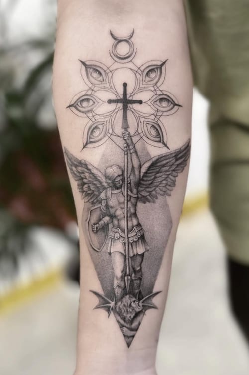 A black and grey tattoo on the forearm depicting an angel holding a cross, with detailed shading and a geometric background — by Jian Liang, los angeles