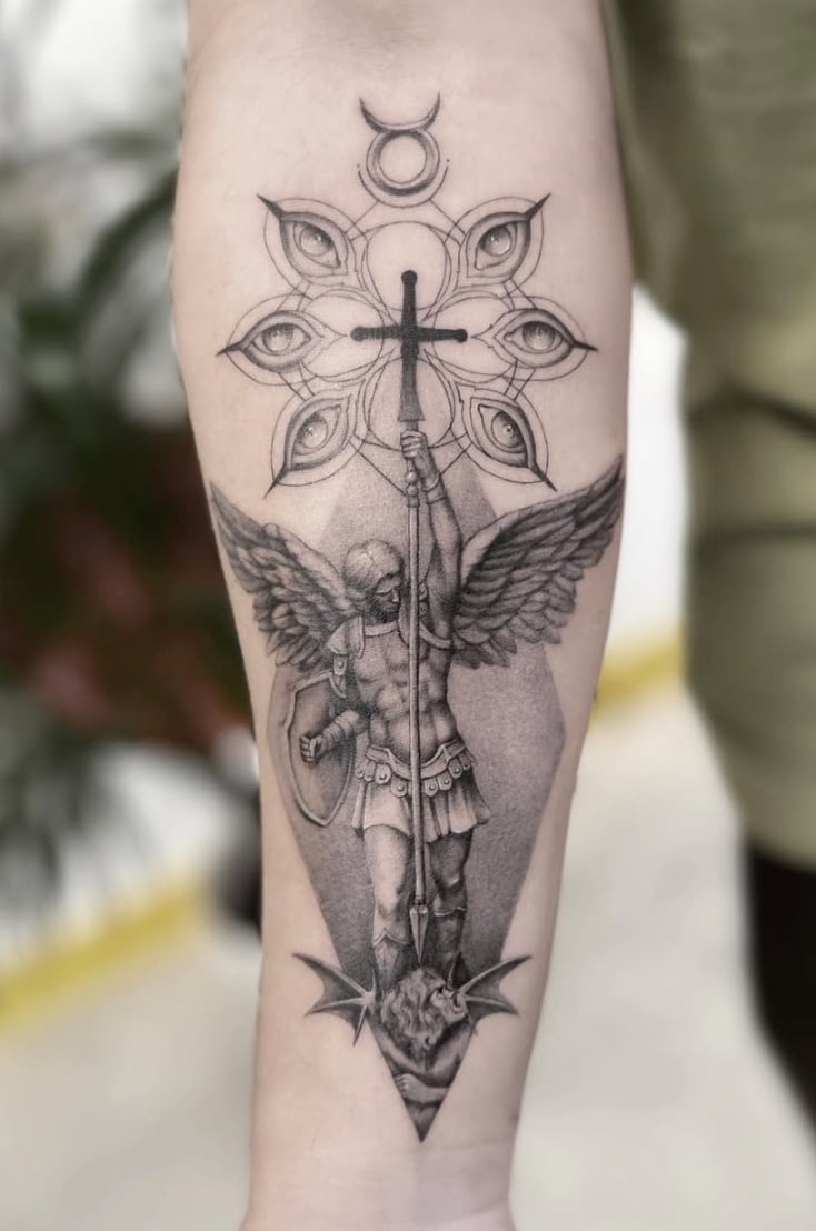 A black and grey tattoo on the forearm depicting an angel holding a cross, with detailed shading and a geometric background — by Jian Liang, los angeles