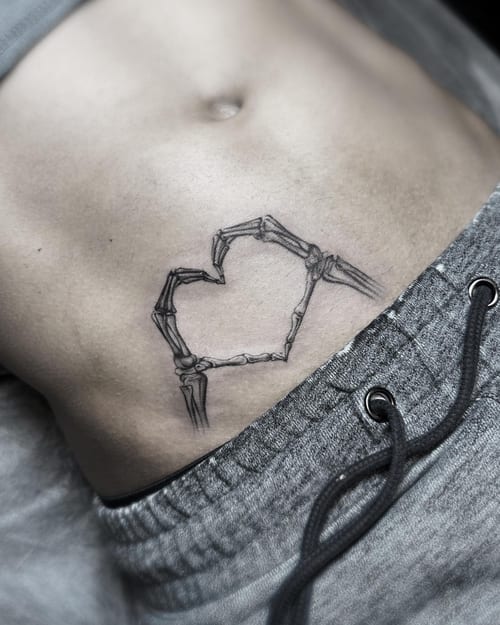 A minimalist blackwork tattoo of skeleton hands forming a heart on the stomach — by Jian Liang, los angeles