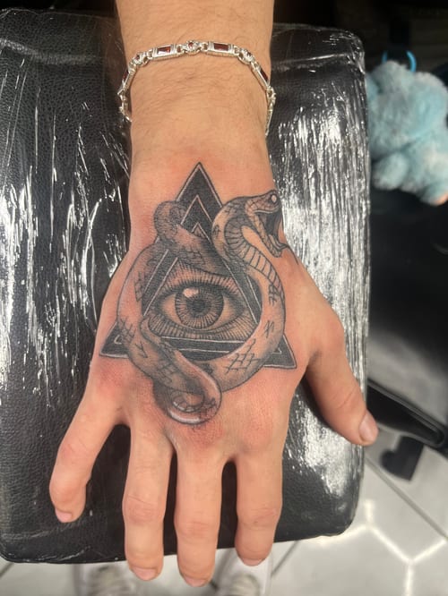 A black and grey tattoo on the hand featuring a geometric eye with a snake wrapped around it, showcasing intricate linework and shading — by Mr._Folks, los angeles