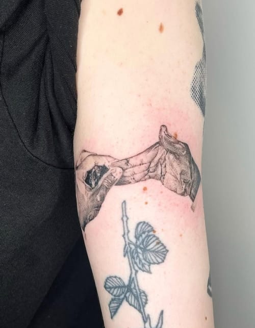 A black and grey realism tattoo on the upper arm depicting two hands reaching towards each other with a floral element below — by Level tattoo, los angeles