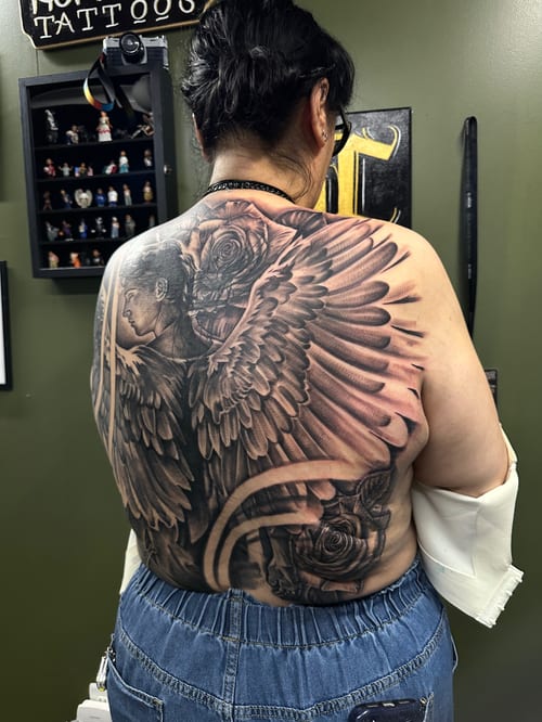A detailed black and grey tattoo of an angel with wings and roses covering the full back, featuring realistic shading and intricate details — by Humilde tattoos, los angeles