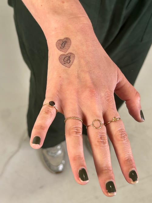 A minimalist black and grey tattoo of two small hearts with initials on the hand — by Brian Kies, los angeles