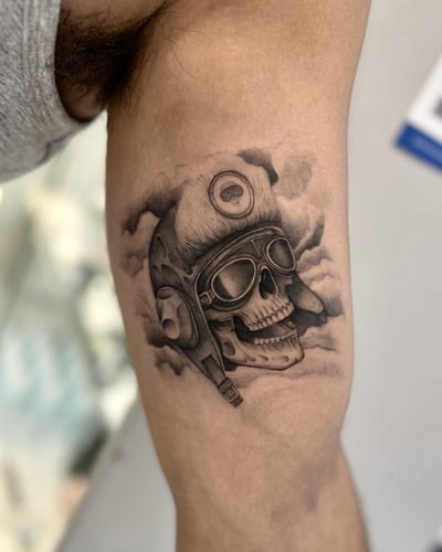 A black and grey realism tattoo of a skull wearing an aviator helmet and goggles on the upper arm, with detailed shading and cloud background — by otaybeatdown, los angeles