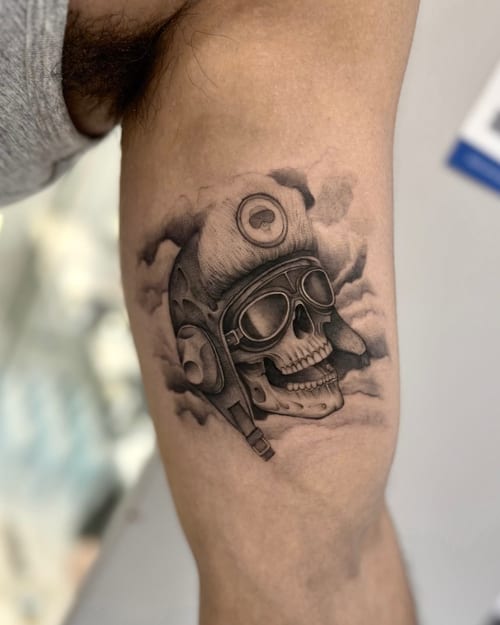 A black and grey realism tattoo of a skull wearing an aviator helmet and goggles on the upper arm, with detailed shading and cloud background — by otaybeatdown, los angeles