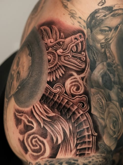 A bold black and grey tattoo featuring an Aztec warrior and a woman's portrait on the shoulder, showcasing intricate details and Chicano style — by Johnny Ayala, los angeles
